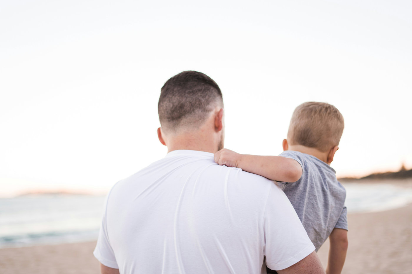A man holds a child on a sandy beach, with gentle waves and a clear blue sky in the background.