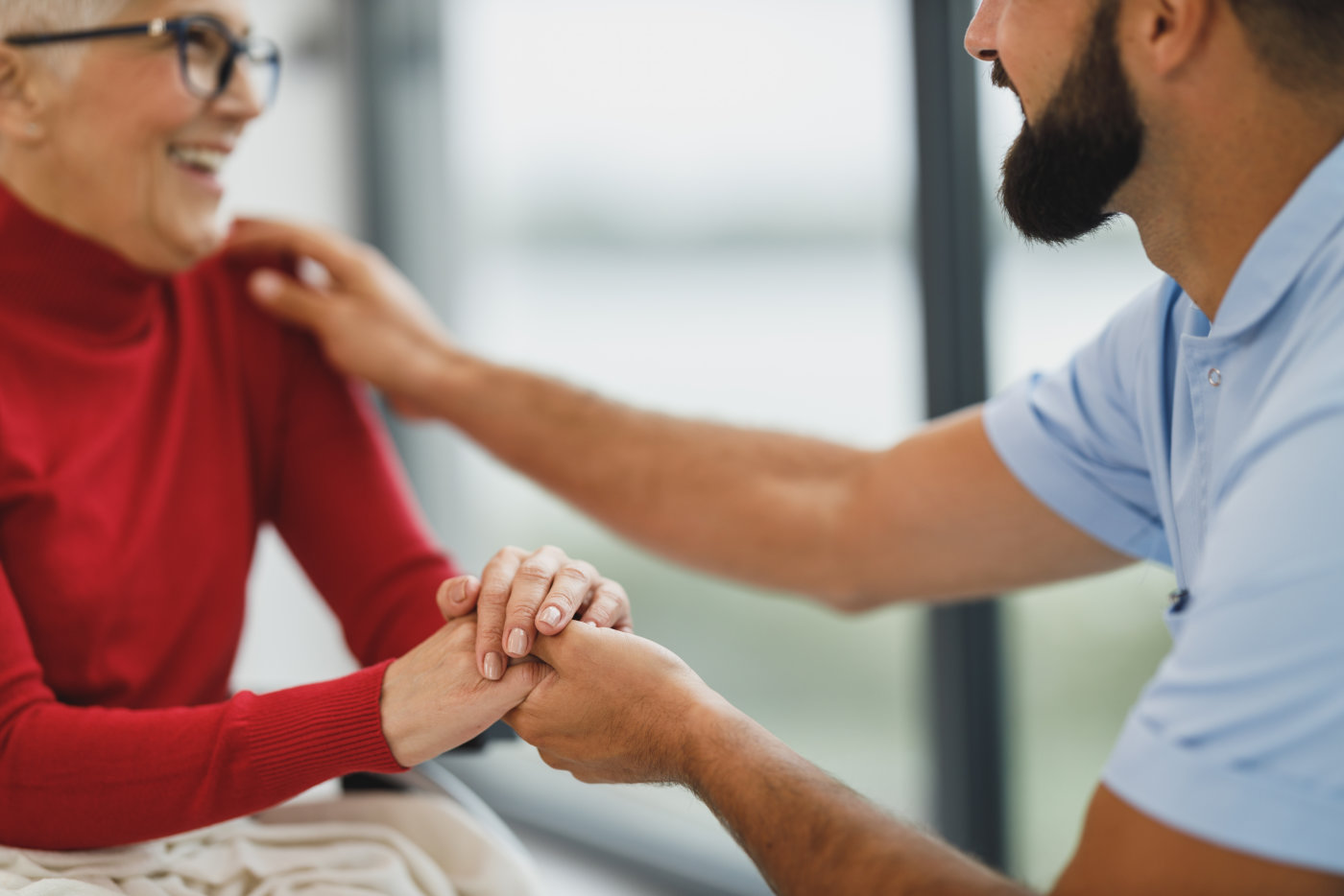 A healthcare worker holds an elderly woman's hand and touches her shoulder in a supportive manner.